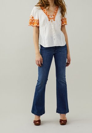 Blouse blanche texturée avec broderie florale orange, manches courtes et détails de boutons, associée à un jean bleu foncé évasé et des sandales marron.
