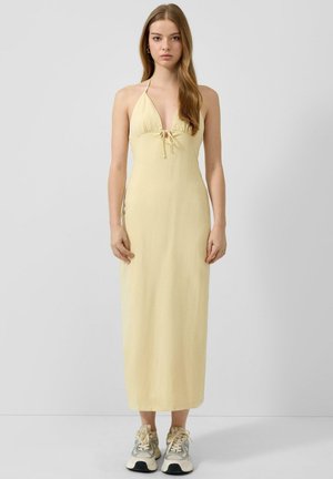 Jeune femme aux longs cheveux portant une robe midi jaune pâle à col licou et des baskets gris-blanc, debout devant un fond gris clair uni.