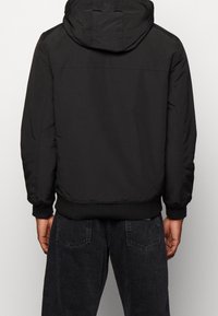 Osoba ubrana w czarną kurtkę z kapturem oraz ribbowymi mankietami i dołem, stojąca tyłem, pokazująca tył kurtki oraz górną część ciemnych dżinsów.