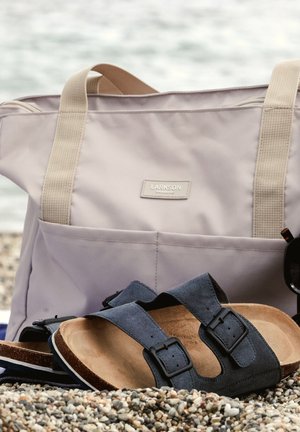 Borsa tote beige con manici in tessuto, sandali in suede blu scuro con fibbie e occhiali da sole su uno sfondo di spiaggia rocciosa.