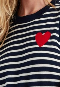 Maglione a righe navy e bianche con un design di cuore rosso sul petto sinistro. Presenta un colletto a girocollo e una texture morbida.
