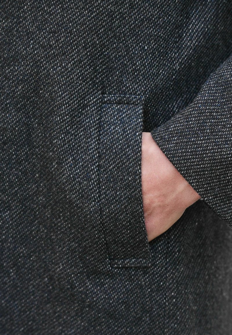 Cappotto in tessuto scuro con una tasca sul petto; mano visibile inserita nella tasca, che mette in mostra il motivo della trama e i dettagli di cucitura.
