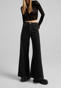 Svarta vida jeans med kontrasterande sömmar, hög midja och en mjuk textur. Bärs tillsammans med en genomskinlig svart randig topp och kraftiga svarta stövlar.