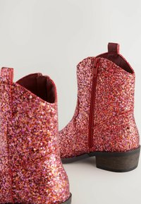 Bottines à paillettes rouges avec une finition brillante, dotées d'une fermeture éclair sur le côté et d'un petit talon en bois, mettant en avant un design texturé et scintillant.