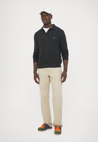 Tamsiai pilkas megztinis su apykakle; smėlio spalvos kelnės su logotipu; spalvingi sportbačiai su oranžiniais, mėlynais, žaliais akcentais; balta kepurė.