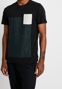 Sort bomulds t-shirt med et teal stribet design på forsiden; har en lille cremefarvet brystlomme og korte ærmer.