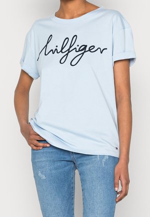 Henkilö, jolla on vaaleansininen T-paita, jonka hihat on kääritty, ja jossa on rintakehän kohdalla "hilfiger"-kirjoitus, sekä keskipesulla olevat revityt farkut.