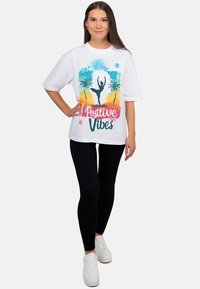 Wit T-shirt met een kleurrijke print van palmbomen en een danser, met de woorden "Positive Vibes". Gecombineerd met zwarte leggings en witte sneakers.
