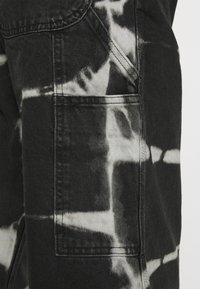 Pantalones de mezclilla negra con un patrón de tie-dye en blanco, que incluyen un bolsillo lateral y detalles de costura en contraste. Textura suave con un tejido visible.