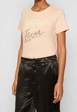 Jasnopomarańczowa koszulka z logo "GUESS" w kolorze żółtym i zdobionymi srebrnymi literami. Okrągły dekolt i krótkie rękawy. W połączeniu z błyszczącymi czarnymi spodniami.