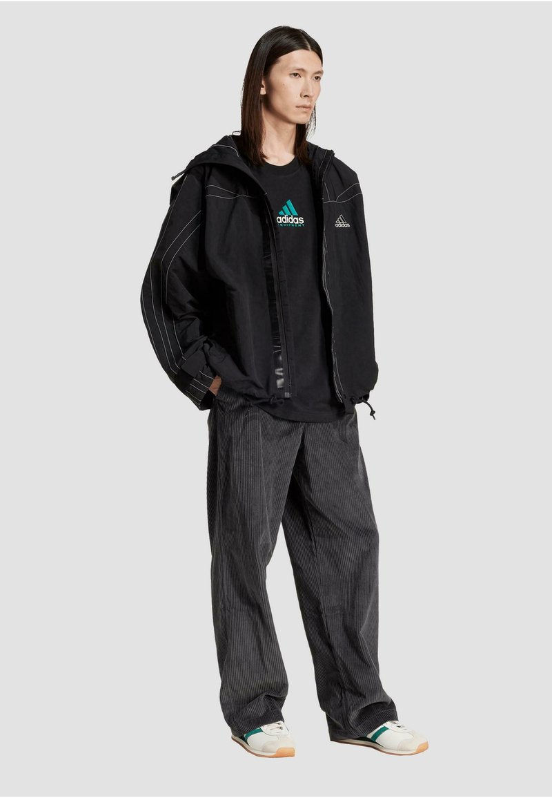 Sort jakke med hvid syning, parret med en sort Adidas t-shirt med et grønt logo og grå corduroy bukser med bred ben. Hvide sneakers.