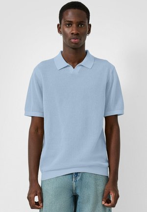 Uomo con polo a maniche corte azzurro chiaro dalla trama particolare e jeans blu, in piedi davanti a uno sfondo bianco semplice.