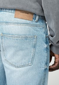 Vaqueros de mezclilla azul claro con un acabado texturizado, que presentan un bolsillo trasero y una etiqueta de piel marrón con la inscripción "EIGHTYFIVE" en la cintura.