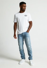 Valkoinen lyhythihainen t-paita mustalla graafisella printillä, yhdessä vaaleansinisten kulutettuja farkkuja ja valkoisten lenkkarien kanssa. Siisti muotoilu, rento istuvuus.