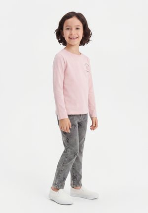 Enfant souriant aux cheveux courts et bruns, debout, portant un t-shirt rose à manches longues, un jean gris avec des motifs de nœuds et des chaussures blanches sans lacets.