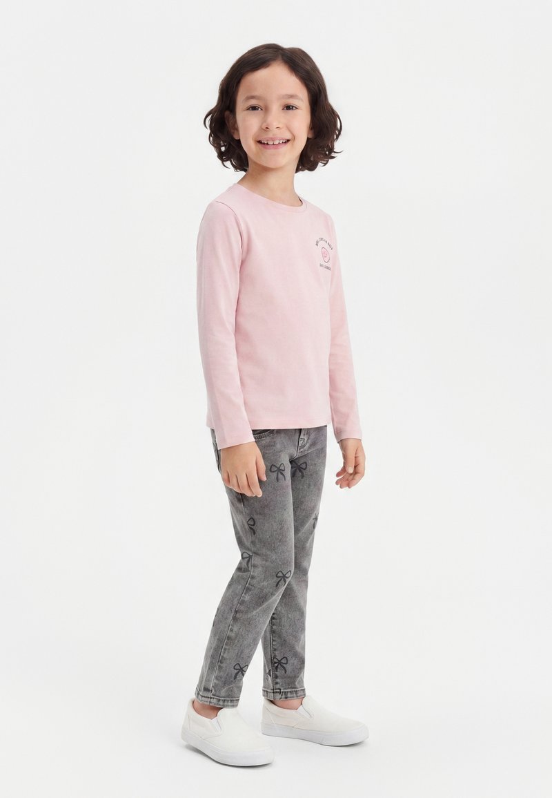 Enfant souriant aux cheveux courts et bruns, debout, portant un t-shirt rose à manches longues, un jean gris avec des motifs de nœuds et des chaussures blanches sans lacets.