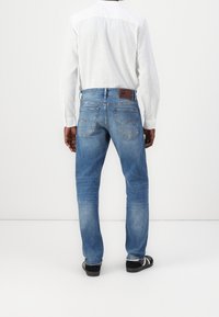 Blå jeans i denim med avslappnad passform, lätt blekning, bakfickor och en brun läderlogotyp på midjebandet. Bärs med en vit skjorta.