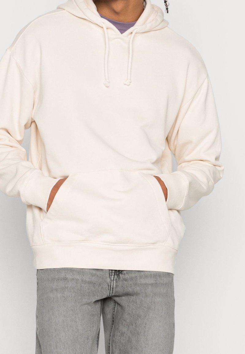 Světlé béžové mikině s kapucí, klokaní kapsou, stahovací šňůrkou a žebrovanými manžetami, doplněné šedými džínami s volnějším střihem a měkkou texturou.