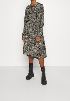 Robe à manches longues avec un imprimé zèbre vert et noir, un col rond avec un détail en forme de goutte, et une jupe fluide. Associée à des bottes noires à lacets.