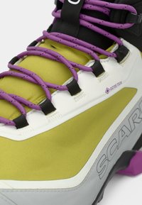 Zapato de senderismo verde y gris con cordones morados, que presenta una parte superior texturizada, ojales reforzados y un logo de Gore-Tex visible.