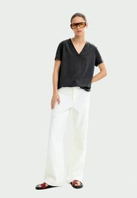 Black v-neck t-shirt, loose fit; white wide-leg pants; black sandals with red accents; large yellow-tinted glasses; neutral background.