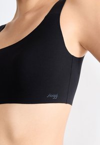 Svart sportbh i slitstark och mjuk tyg. Har breda axelband och låg skärning i halsen. "sloggi"-logotyp i ljusblått nära fållen.