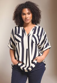 Blusa de manga corta con rayas en azul marino y crema, cuello en V, dos bolsillos en el pecho y corte holgado. Llevada con unos vaqueros azul oscuro. Textura de tela suave.