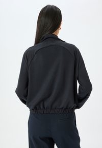 Svart lättviktsjacka med krage, elastisk midja och slät textur, med en subtil lyster och avslappnad passform.