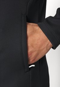 Zwarte sporttop met een zijzak en een witte koord. Glad stoffen textuur, aansluitend ontwerp en versterkte stikseldetails.