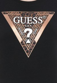 GUESS-logo på sort stof med leopardmønster-triangel, rosaguld studs og teksten "WASHED JEANS" omkring et stort hvidt spørgsmålstegn.