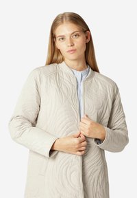 Chaqueta acolchada beige con cuello redondo, cremallera delantera y patrones en espiral texturizados, llevada sobre una camisa azul claro.