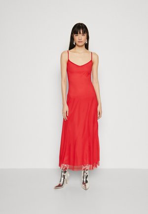 Red satin maxi dress with thin straps, featuring a semi-sheer hem. Model wears silver metallic pointed-toe boots. Background is plain.