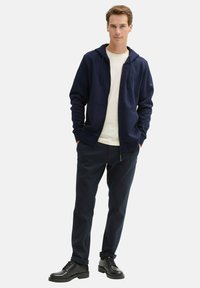 Marineblauwe rits-hoodie met koorden, gedragen over een wit shirt. Gecombineerd met donkere broek en zwarte leren schoenen. Gladde stoftextuur.