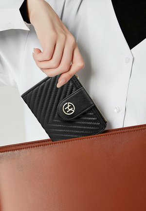 Billetera de cuero negra con acolchado en forma de chevron, herrajes dorados y cierre a presión, sostenida por encima de un bolso plano marrón.