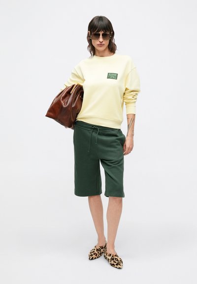 Sweat-shirt jaune avec logo vert, short vert et ballerines imprimé léopard. Porte un sac marron, des lunettes de soleil. Mannequin debout sur fond blanc.