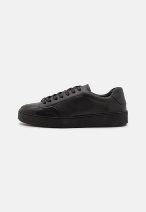 Zapatillas de cuero negro con puntera redondeada, suela plana y cordones tradicionales. Presentan una textura suave y un sutil detalle en los paneles laterales.