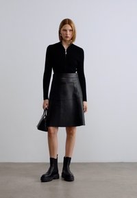 Μαύρο πουλόβερ με φερμουάρ, μαύρη δερμάτινη φούστα μέχρι το γόνατο και χοντρά μαύρα μποτάκια. Το μοντέλο κρατά μια μικρή μαύρη τσάντα χειρός. Μινιμαλιστικός σχεδιασμός.