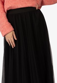 Gonna nera in tulle con pieghe e superficie textured, dotata di waistband elastico nero. Indossata con un maglione lavorato a maglia color corallo.