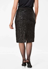 Schwarzer Pailletten-Bleistiftrock mit Schlitz hinten, ausgestattet mit einer glatten Textur und figurbetontem Schnitt. Das Model trägt schwarze High Heels und Strumpfhose.