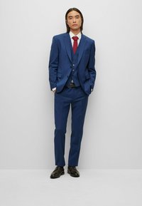 Blauer dreiteiliger Anzug mit einer Zwei-Knopf-Jacke, passendem Weste und glattem Hosenbund. Weißes Hemd und rote Krawatte; schwarze Schuhe vervollständigen den Look.