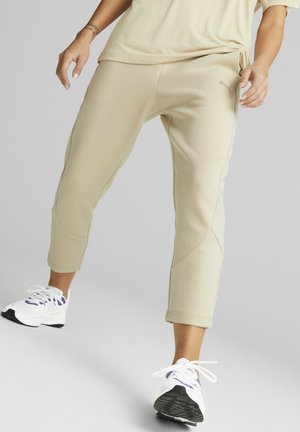 Osoba ubrana w beżowe, krótkie spodnie dresowe i białe trampki, stawiająca krok do przodu na jednolitym szarym tle.