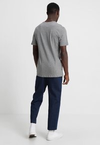 Šedé bavlněné tričko s kulatým výstřihem, v kombinaci s tmavě modrými kalhotami a bílými teniskami, z pohledu zezadu. Jednoduchý design, bez viditelných vzorů.