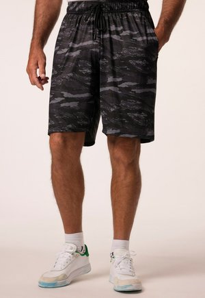 Ein Mann trägt schwarze und graue Shorts mit Tarnmuster, kombiniert mit weißen Sneakers und weißen Socken, und steht vor einem schlichten Hintergrund.