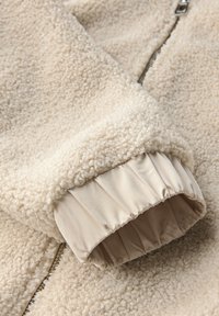 Primo piano di una manica di una giacca in pile beige con polsino in tessuto elastico e cerniera argentata visibile su un materiale in lana lavorato.