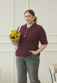 Pull en tricot bourgogne à manches courtes avec un col à patte et trois boutons, associé à un jean gris et un sac tissé contenant des tournesols.