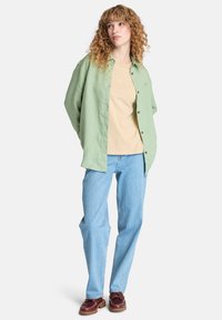 Camicia a maniche lunghe verde chiaro sopra una maglietta beige, abbinata a jeans azzurri e scarpe marroni con suola spessa. Outfi casual a strati.