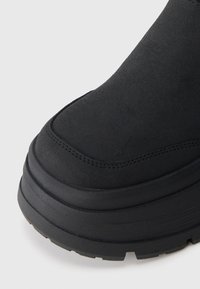 Fekete velúr csizma orra vastag gumi talppal és dupla varrási részletekkel, fehér háttérrel.