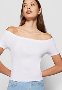 Valge off-the-shoulder topp lühikeste varrukatega, kitsas lõige, soonik tekstuur ja lühike pikkus, koos helesiniste teksadega.