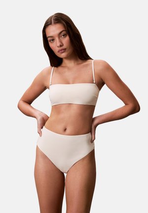 Femme aux longs cheveux bruns portant un haut de bikini bandeau beige et un bas de bikini taille haute sur un fond clair uni.