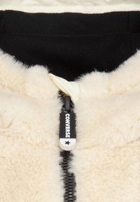 Kremowa polarowa kurtka o miękkiej fakturze, z czarnym zamkiem błyskawicznym i markowym uchwytem, częściowo podszyta czarnym materiałem.